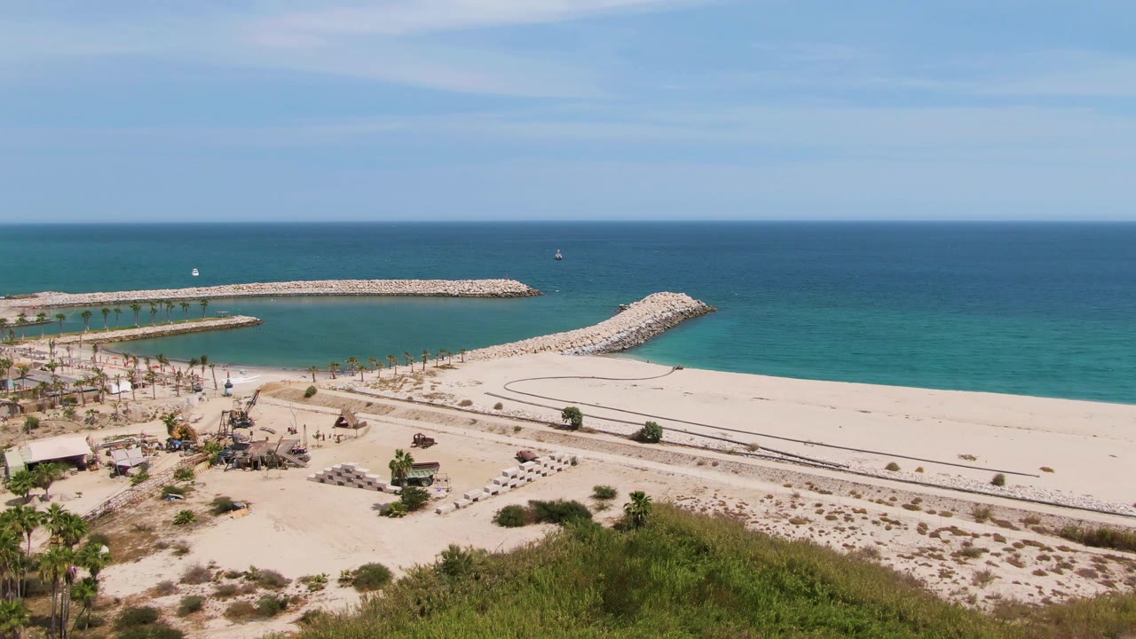 Wide aerial view of sandy beach, turquoise ocean, rocky breakwater, palms, and dune greenery.