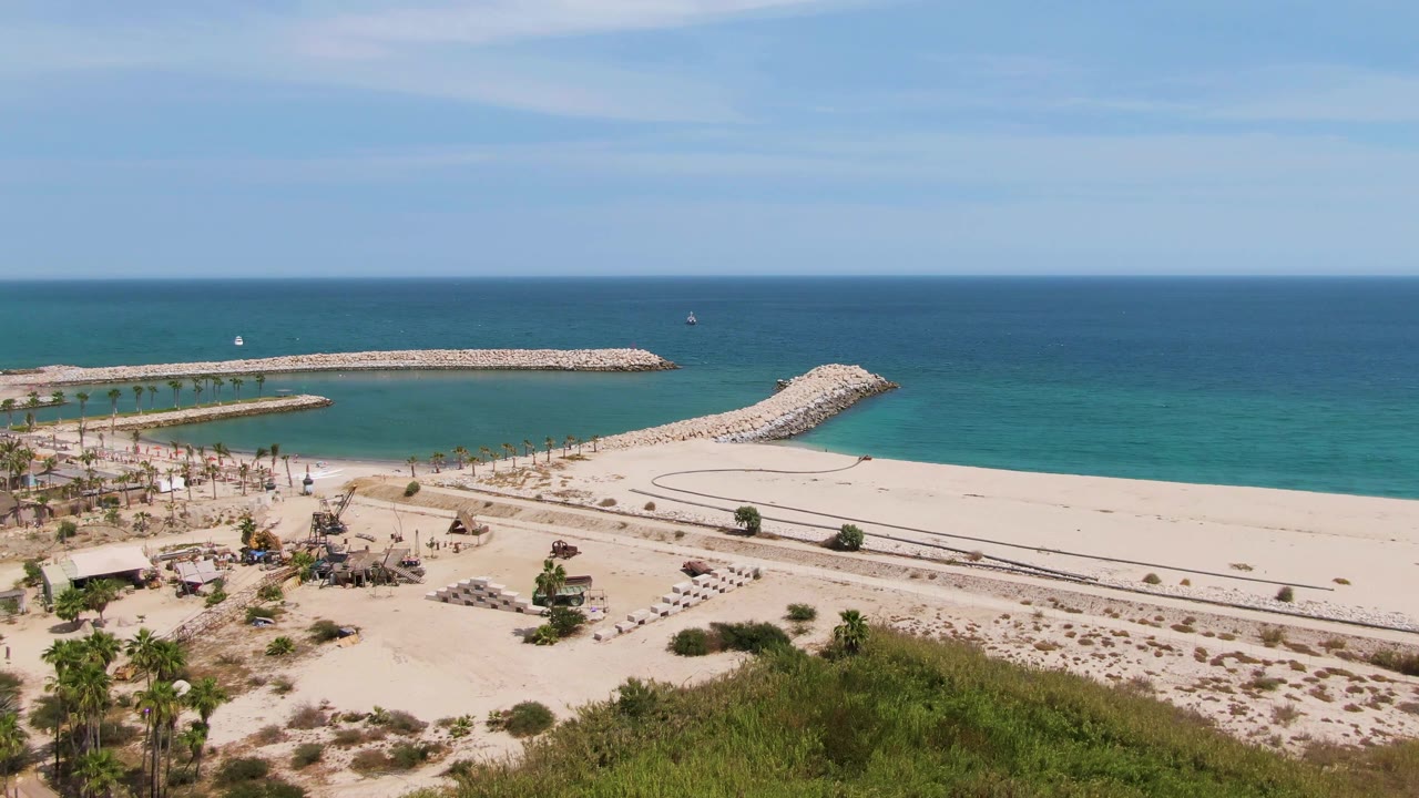 At 0:01, the Puerto Los Cabos Beach aerial view keeps the breakwater and horizon centered while palms, beach paths, rows of lounge chairs, and no visible people remain in frame.