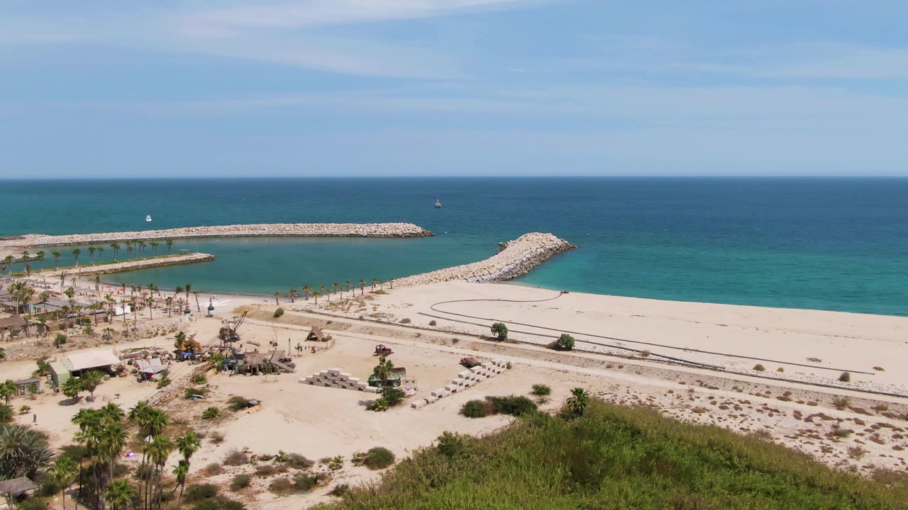 At 0:02, Puerto Los Cabos Beach is shown with more foreground palms and shrubs visible below the open sand, plus the rocky breakwater, blue ocean, clear sky, and no visible people.