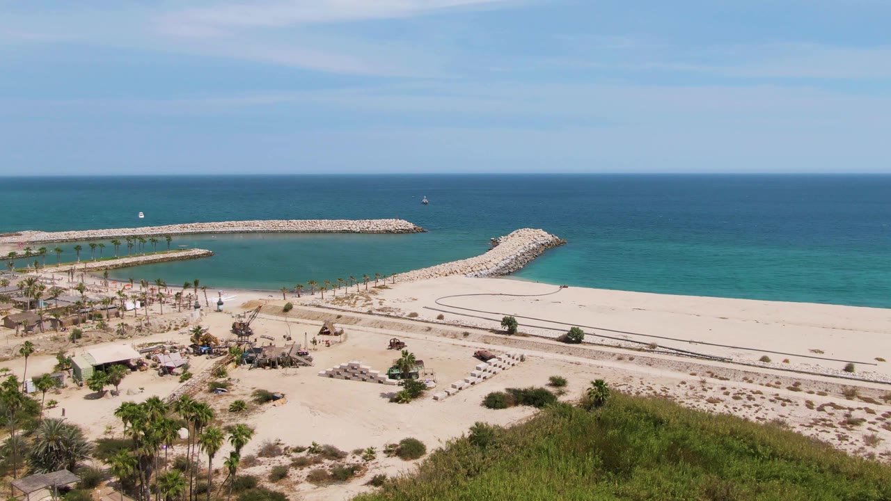 At 0:03, the aerial view of Puerto Los Cabos Beach shows the smaller inlet jetty at left, the long rocky breakwater, foreground palms and shrubs, bright sand, and no visible people.