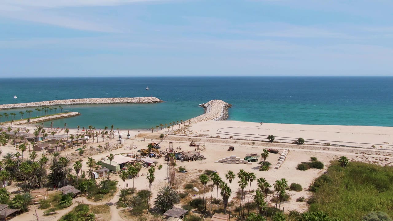 Puerto Los Cabos Beach from above, showing clustered palms and hut roofs at lower left, broad pale sand through the center, calm blue-green water, and rocky breakwaters on the horizon line, with no visible people.