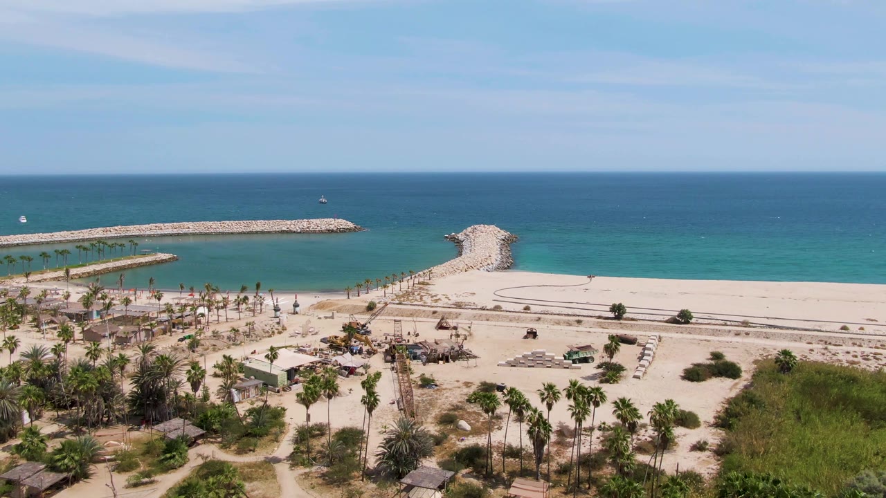 Elevated view of Puerto Los Cabos Beach with the palm grove spread across the lower frame, light sand and sparse beach fixtures in the middle, and a rocky breakwater separating calm turquoise water from the open ocean, with no visible people.