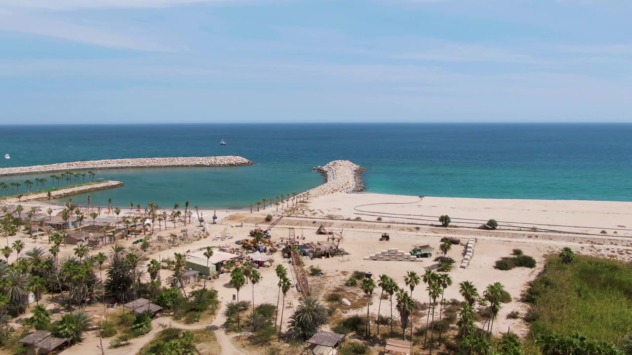 Puerto Los Cabos Beach aerial frame with palms and huts low in view, a mostly empty sandy expanse and road behind them, turquoise nearshore water to the left, and a long stone breakwater running across the ocean, with no visible people.