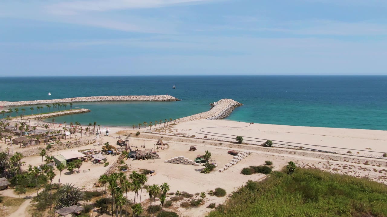 At 0:00, Puerto Los Cabos Beach appears from above with palm trees and green scrub in the foreground, pale sand and curved paths below, turquoise ocean and stone breakwaters beyond, and no visible people.