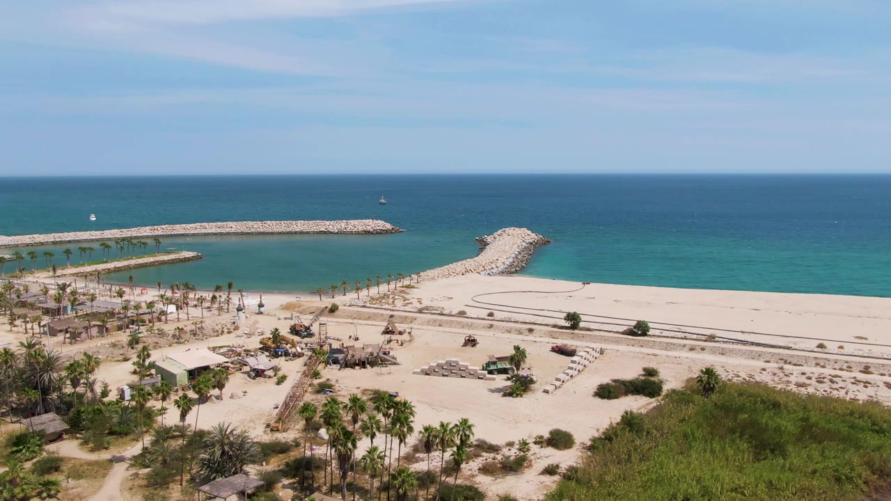 At 0:02, Puerto Los Cabos Beach is seen from an elevated drone angle with the stone jetty crossing the water, a rounded breakwater to the right, beach palms below, and no visible people.