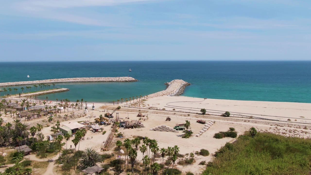 Aerial shoreline view with sand, palms, turquoise sea, and breakwater under clear sky.