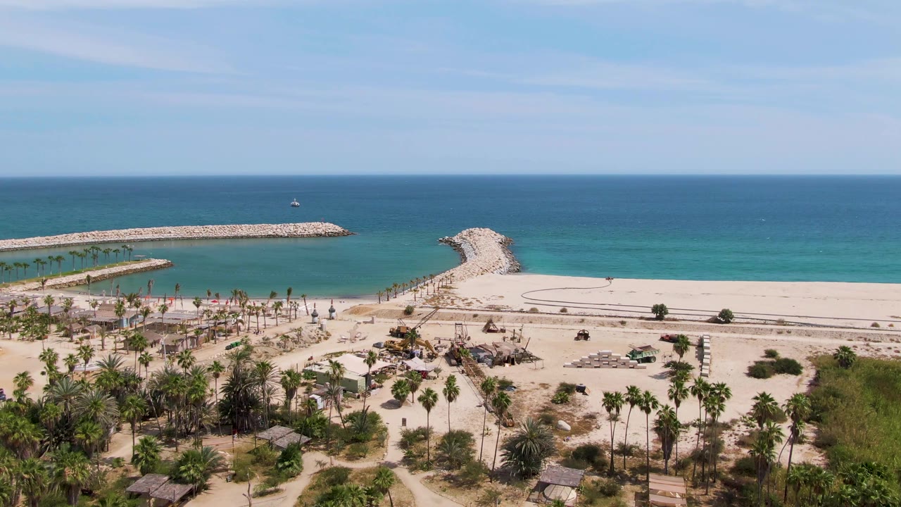 Aerial frame over Puerto Los Cabos Beach showing palms and low beach structures in the foreground, a road beside the sand, turquoise ocean, rocky breakwaters, and no visible people.