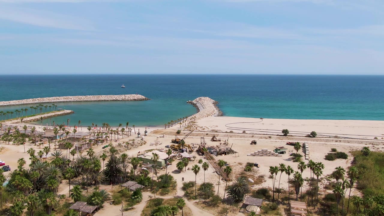 Aerial frame at Puerto Los Cabos Beach with the road and small vehicles crossing the sandy beachfront, palm trees below, breakwaters extending into turquoise water, and no visible people.