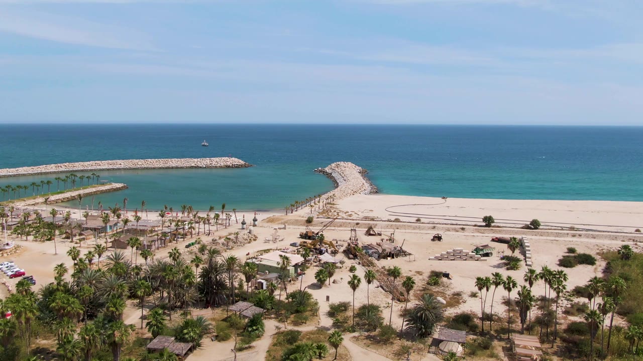 Aerial frame over Puerto Los Cabos Beach with more parked cars visible near the palms, the beachfront road across the sand, calm turquoise water, rocky piers, and no visible people.