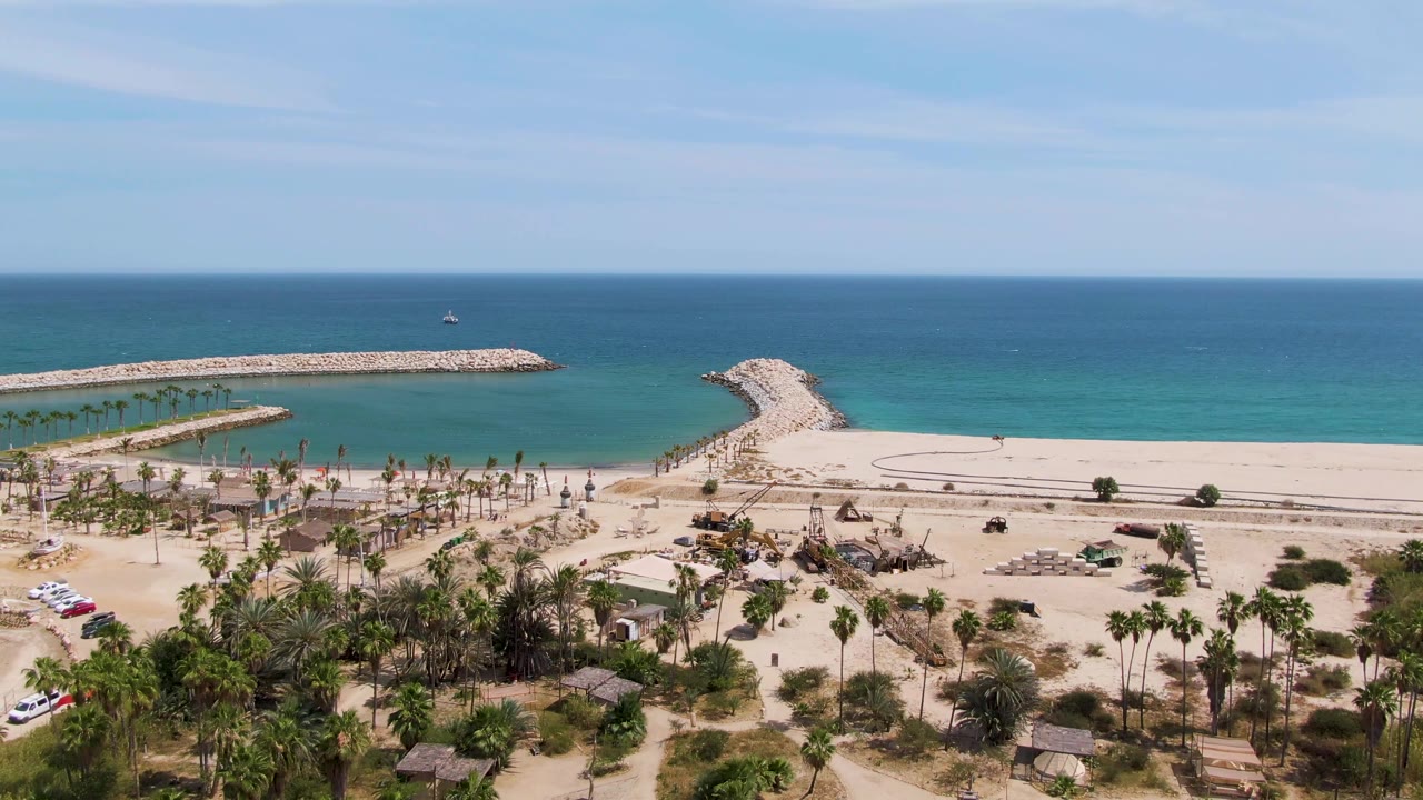 Aerial beach road scene with cars at left, palms below, sand, ocean, and breakwaters.