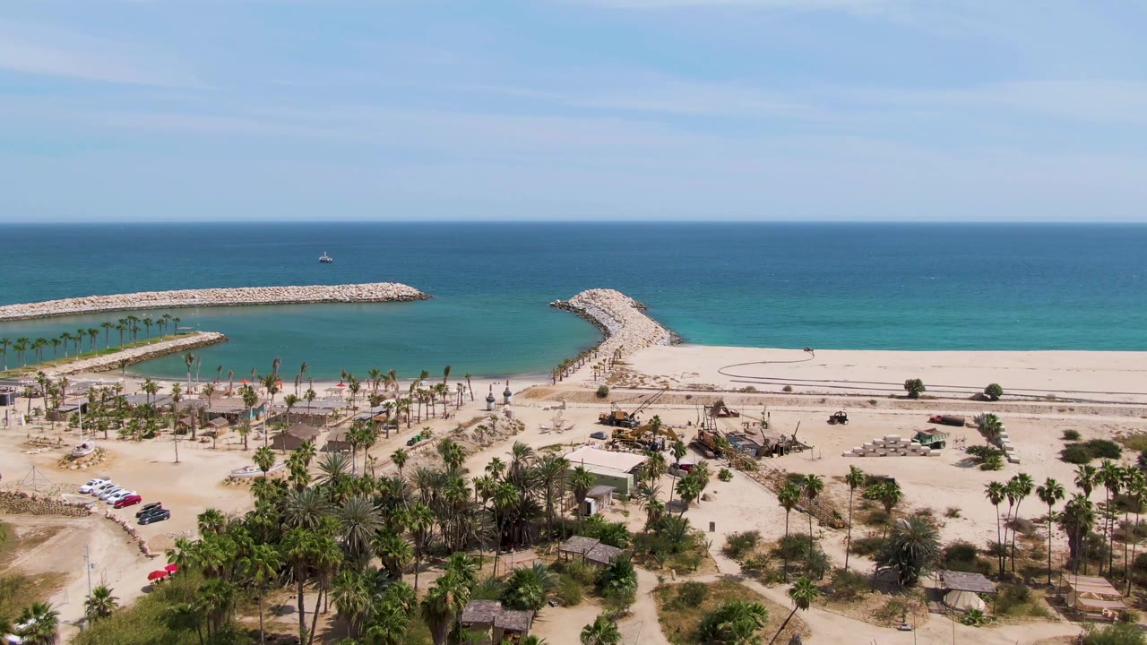 Aerial frame over Puerto Los Cabos Beach showing sandy parking, palm trees, a red car near the lower left area, stone breakwaters, turquoise ocean, and no visible people.