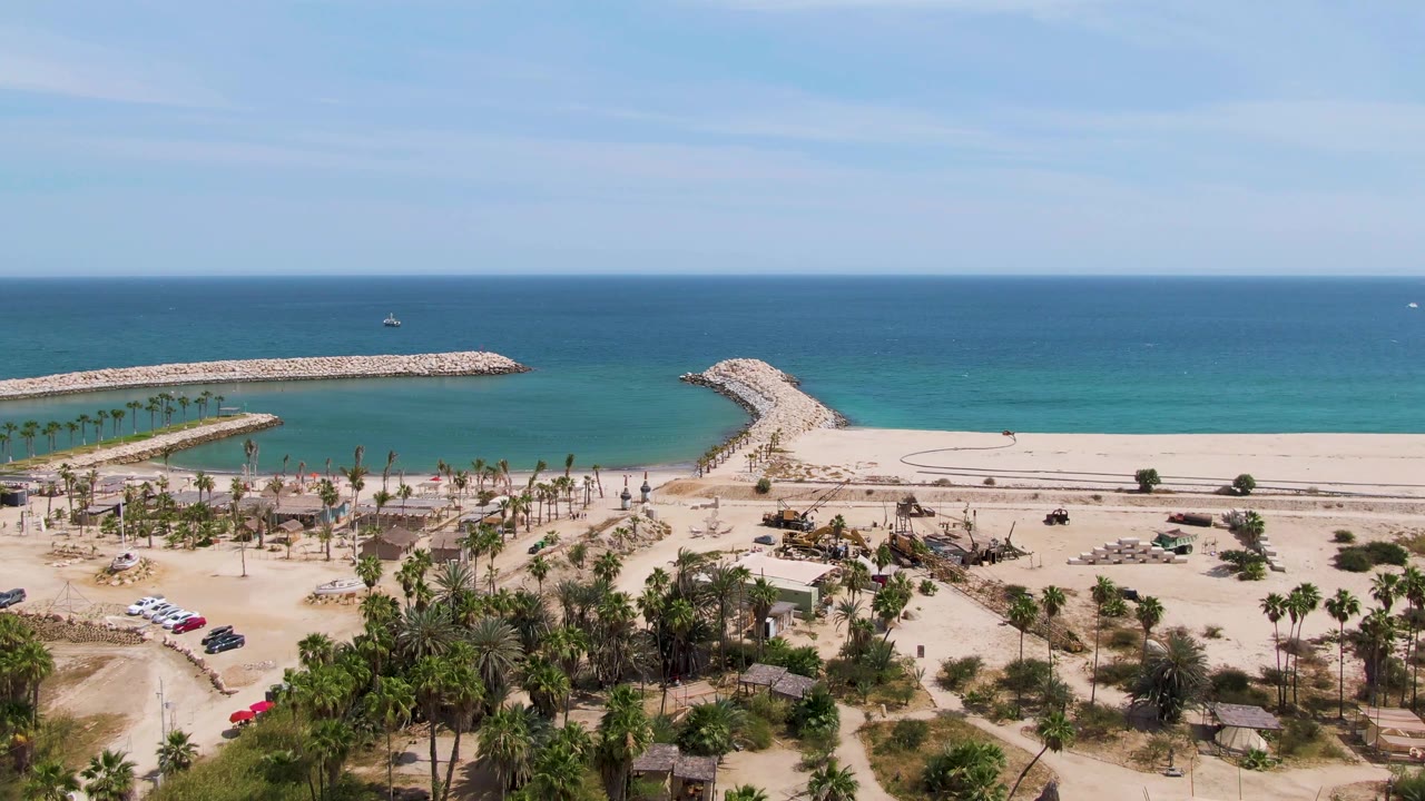 Aerial view at Puerto Los Cabos Beach with foreground palms, scattered parked vehicles, the red car along the sandy lot, pale beach, curved breakwater, and no visible people.