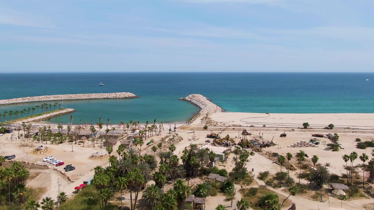 Puerto Los Cabos Beach aerial frame with the red car shifted slightly through the sandy parking area, palm clusters below, two stone breakwaters ahead, blue water, and no visible people.