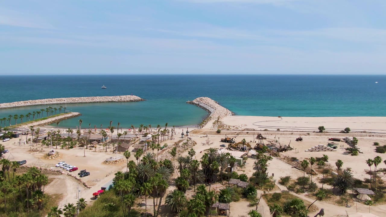 Wide aerial beach scene with palms, vehicles, breakwaters, sand, and ocean.
