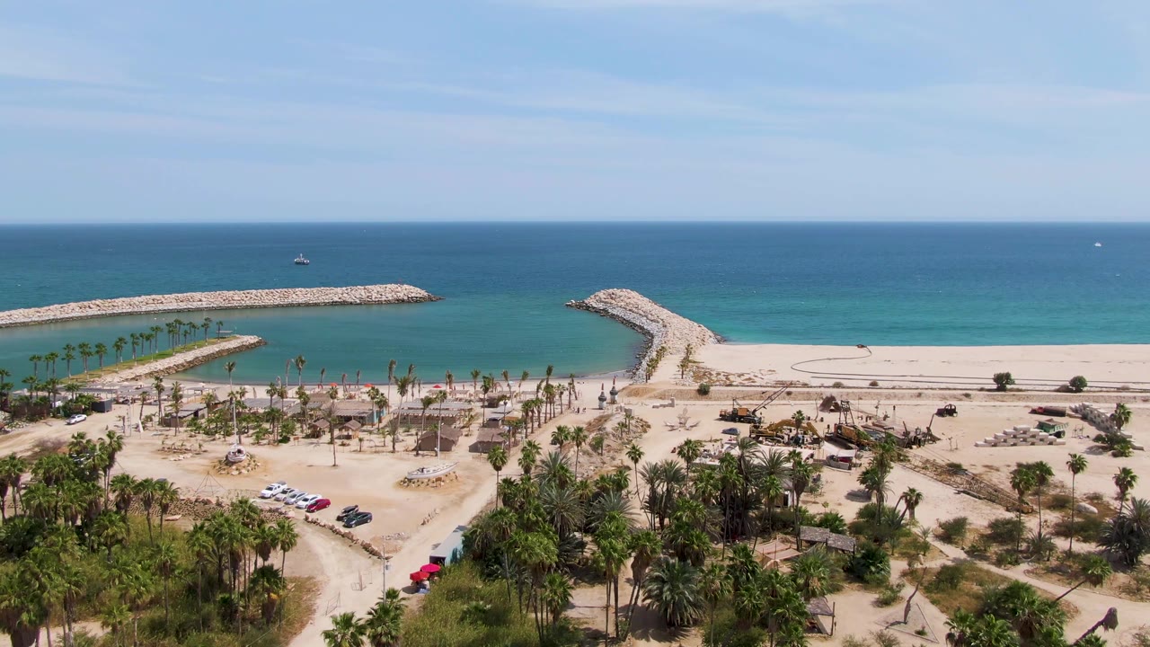 At 0:00, Puerto Los Cabos Beach is seen from above with a sandy parking area, cars, palms, clear turquoise water, two rocky breakwaters, and no visible people.