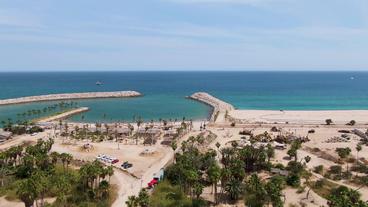 At 0:01, the aerial view over Puerto Los Cabos Beach keeps the sandy lot and palm grove in the foreground with a beach access path, breakwaters, turquoise ocean, and no visible people.