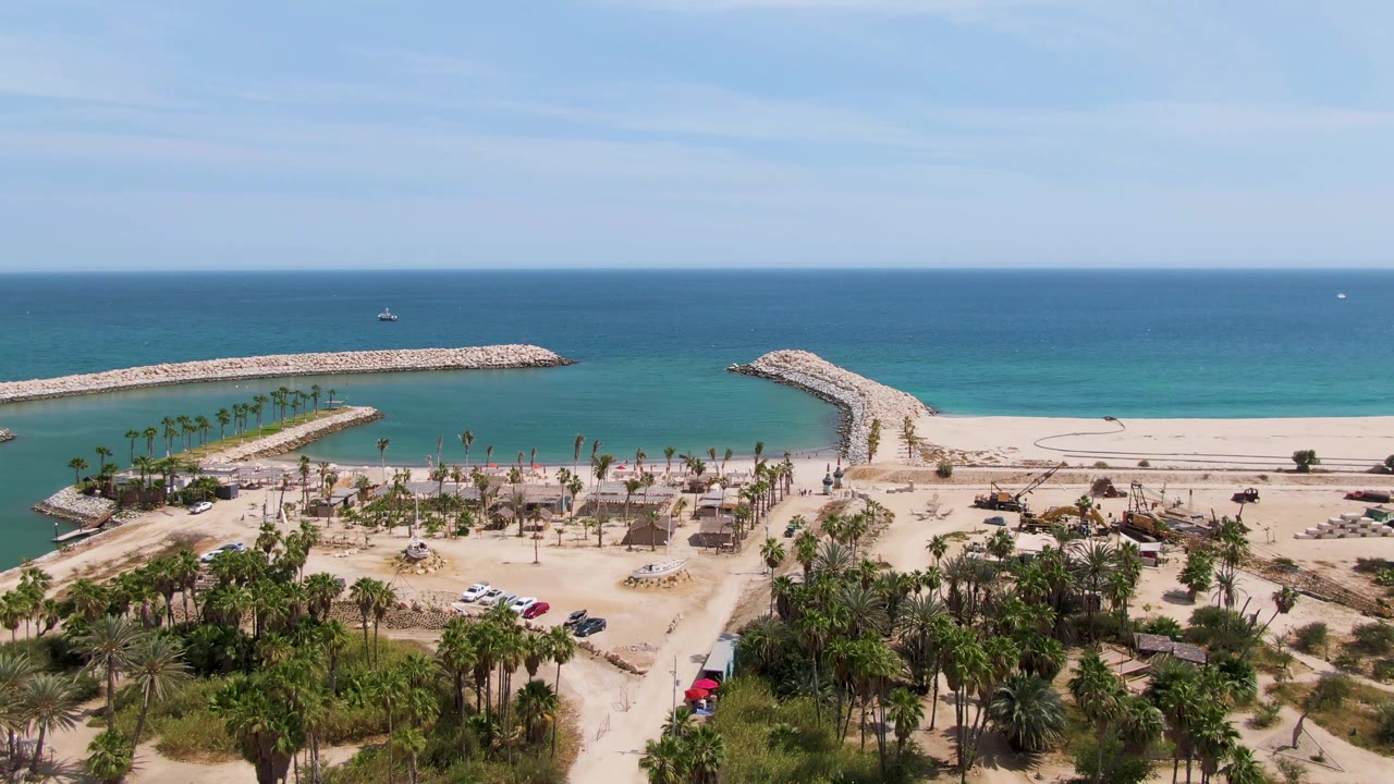 Aerial coastal view with palms, sandy parking, rocky breakwaters, and calm blue water.