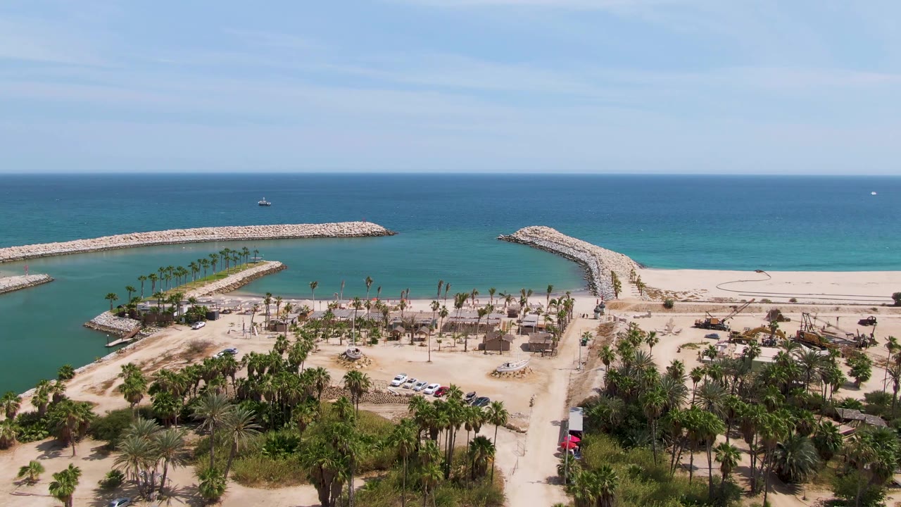 Aerial frame over Puerto Los Cabos Beach showing rocky breakwaters, sandy shore, palm trees, parked vehicles, blue ocean, and no visible people at 0:00.