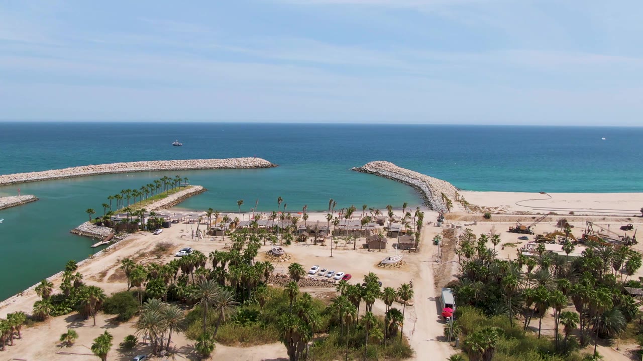 Puerto Los Cabos Beach appears from above with two rocky breakwaters, smooth turquoise shallows, sandy beachfront, palms, scattered vehicles, and no visible people at 0:02.