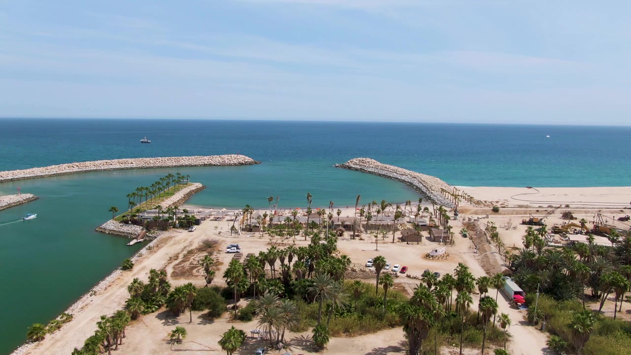 Aerial frame over Puerto Los Cabos Beach showing a sandy channel between rock breakwaters, turquoise water, palm trees, parked vehicles, open ocean, and no visible people.