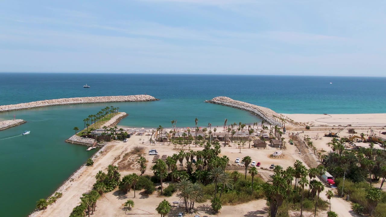 Drone-style aerial view at Puerto Los Cabos Beach with a small boat beginning to cross the green-blue channel beside sand, palms, parked vehicles, breakwaters, and no visible people.
