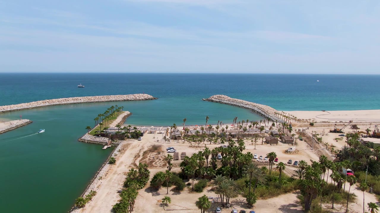 Aerial frame over Puerto Los Cabos Beach with the boat and wake visible in the channel, palms and sand below, rock breakwaters enclosing the water, open ocean beyond, and no visible people.