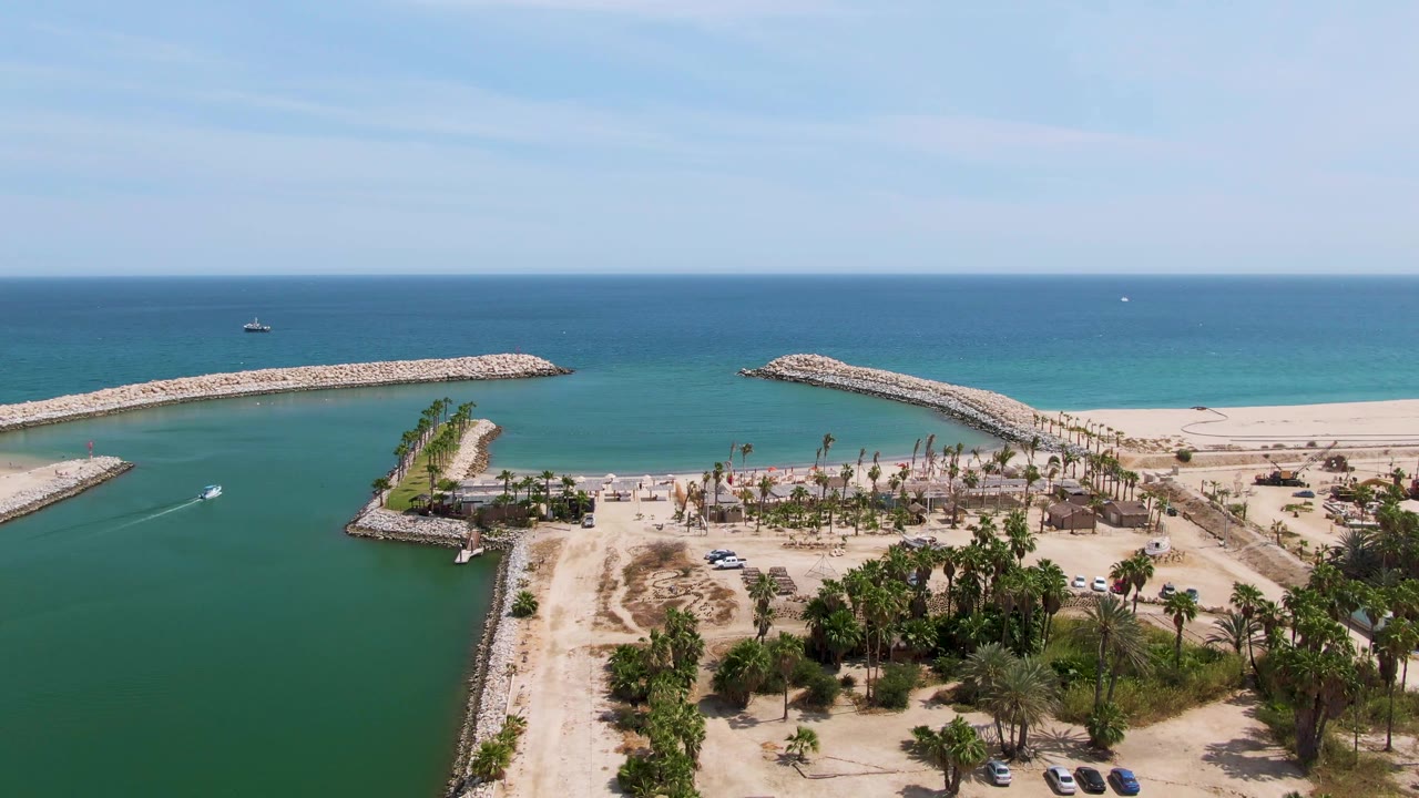 Aerial view of a coastal channel, rock breakwaters, sandy beach, palms, cars, and blue ocean.