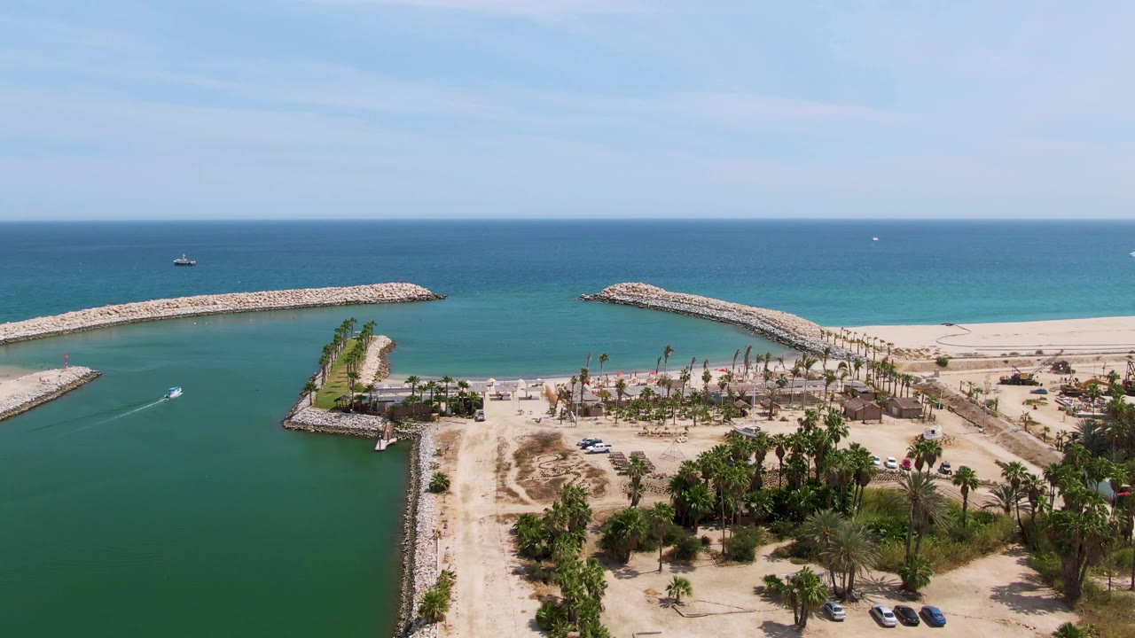 Second frame at Puerto Los Cabos Beach with the channel filling the foreground, a small boat wake on the left water, rock breakwaters, beach palms, cars, and no visible people.