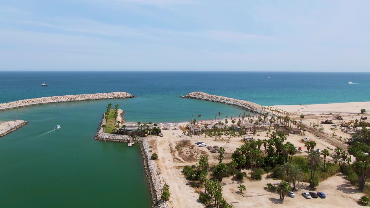 Fourth frame at Puerto Los Cabos Beach with the left breakwater extending into the channel, a small white boat track on the water, sandy construction-like ground, palms, cars, and no visible people.