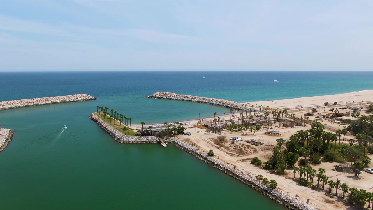 Aerial frame at Puerto Los Cabos Beach showing a small boat entering green-turquoise harbor water between rocky breakwaters, with sandy beach, palms, open ocean, and no visible people.