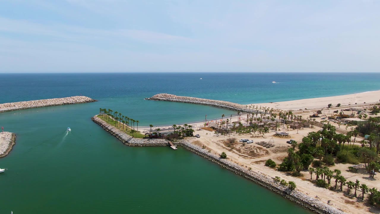 Puerto Los Cabos Beach aerial view with the small boat farther into the harbor channel, a white wake behind it, rocky breakwaters, docked boats at left, sand, palms, and no visible people.