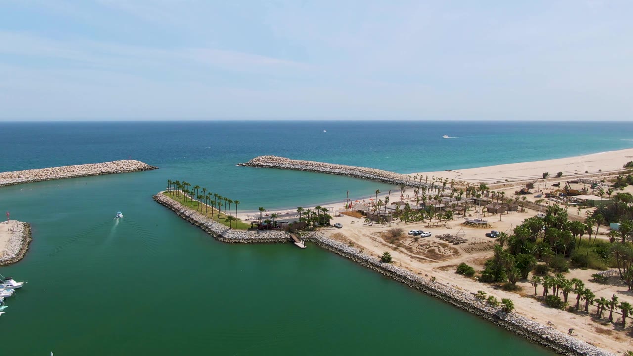 Aerial frame over Puerto Los Cabos Beach with the harbor channel centered, the small boat and wake visible on the water, sandy shoreline and palms behind the breakwaters, and no visible people.