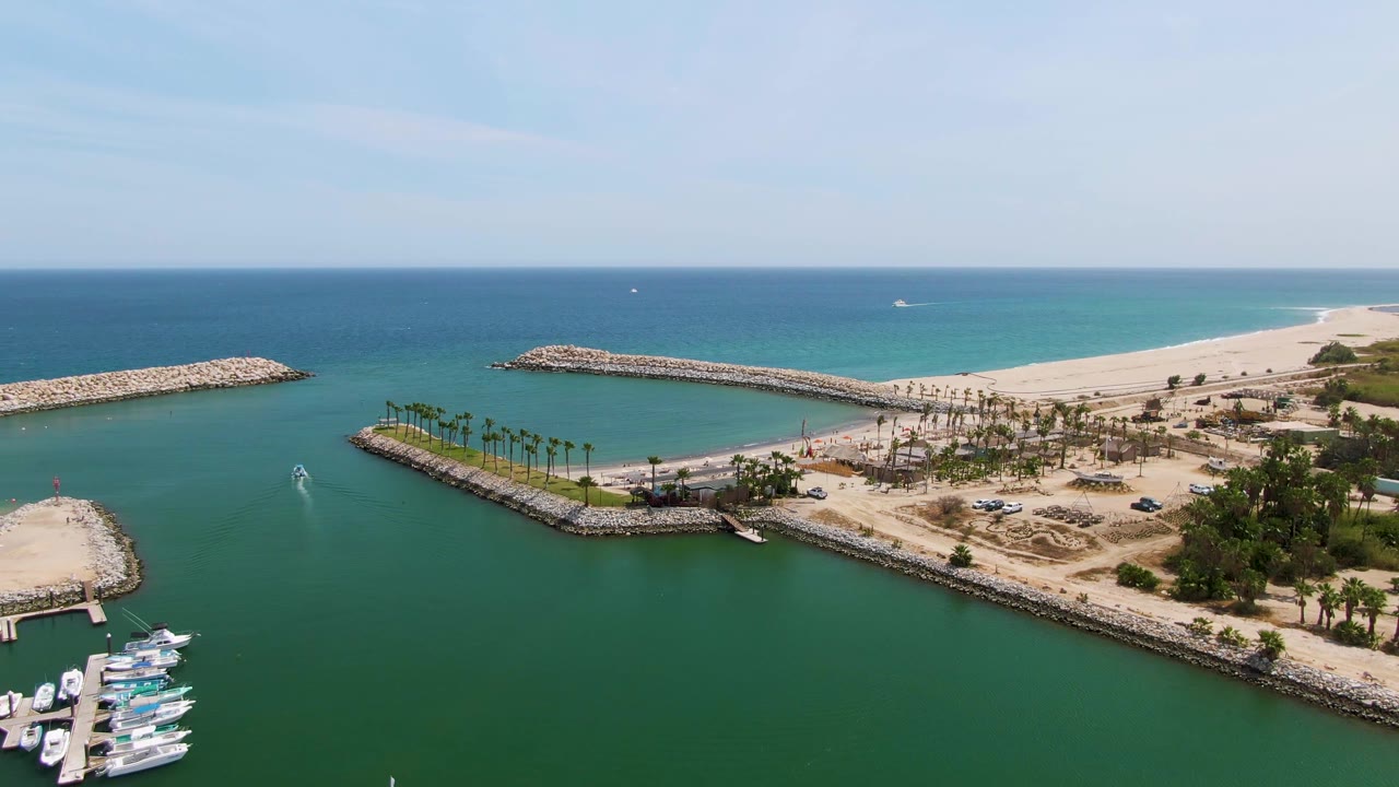 Opening aerial frame over Puerto Los Cabos Beach showing green marina water, docked boats along the lower edge, rocky breakwaters, palm trees, sandy beach, blue ocean, and no visible people.