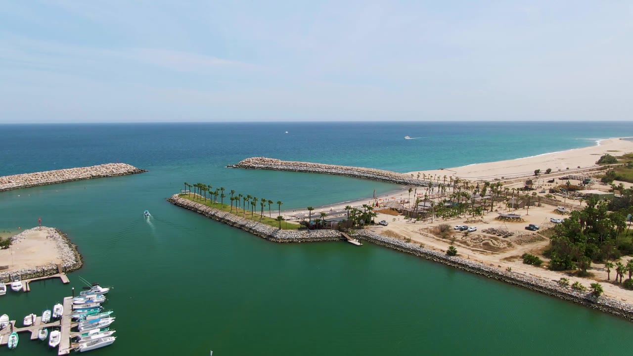 Second aerial frame at Puerto Los Cabos Beach with the docked boats lower right, a palm-lined strip near the marina, a small wake in the green water, sandy beach beyond, and no visible people.