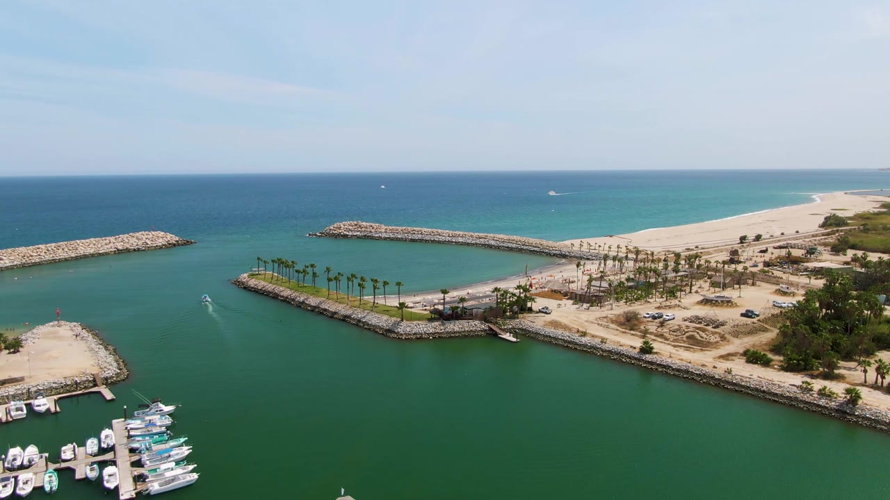 Aerial coastal marina view with green water, boats, palms, sand, and breakwaters.