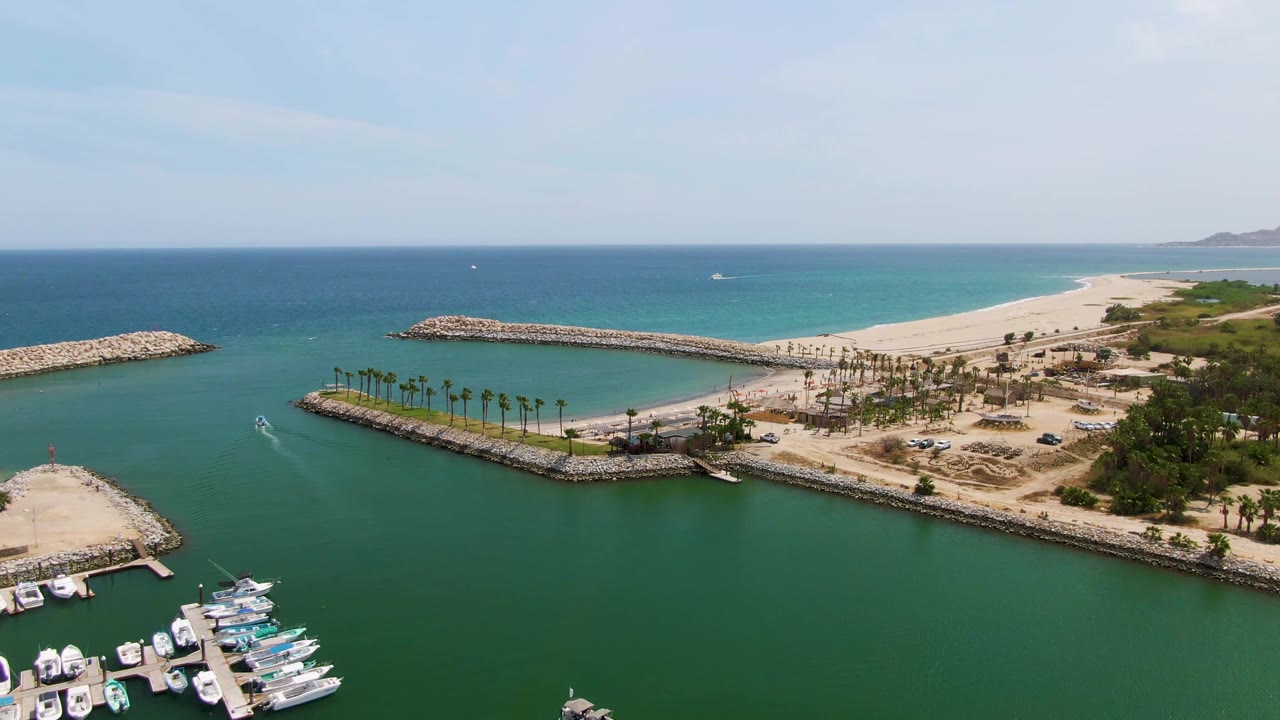 Second frame of Puerto Los Cabos Beach harbor, with the white boat wake slightly farther through the green water, yachts clustered at the lower edge, breakwaters enclosing the marina, and no visible people.