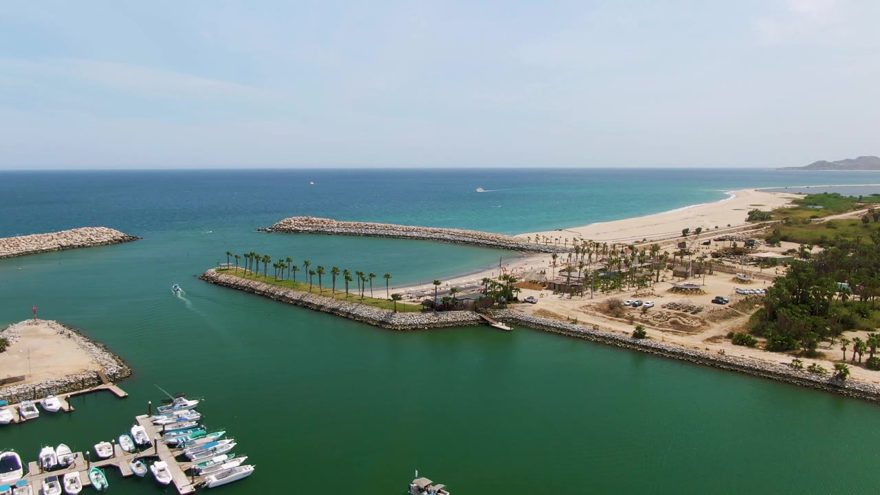 Third frame over Puerto Los Cabos Beach, showing the boat wake crossing the harbor water, the docked yachts partly cropped at bottom, a palm-lined breakwater, sandy shore, blue ocean, and no visible people.
