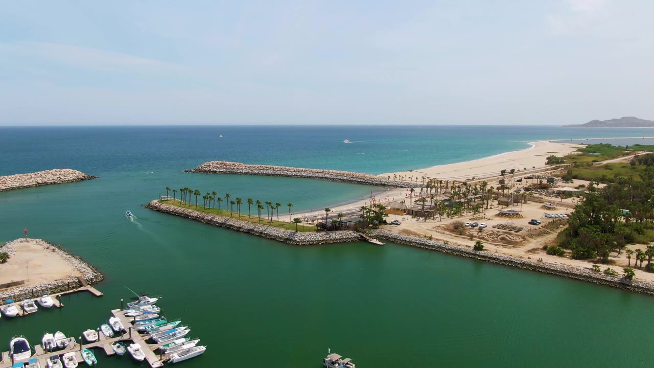 Fourth frame looking across Puerto Los Cabos Beach harbor, with the boat wake still visible in the green basin, yacht slips near the lower left, rock breakwaters framing the water, beach and palms beyond, and no visible people.