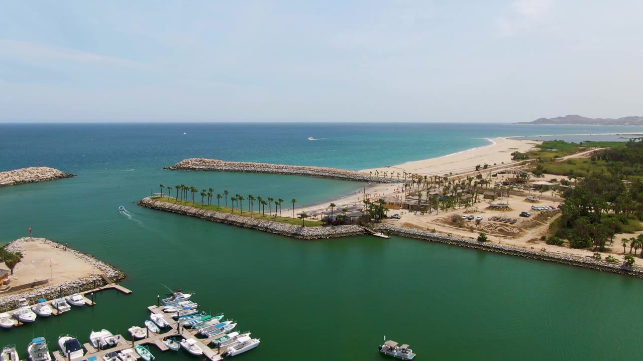 Aerial frame over Puerto Los Cabos Beach showing docked marina boats at the lower edge, green water, a palm-lined breakwater, sandy shore, blue ocean, distant hills, and no visible people.