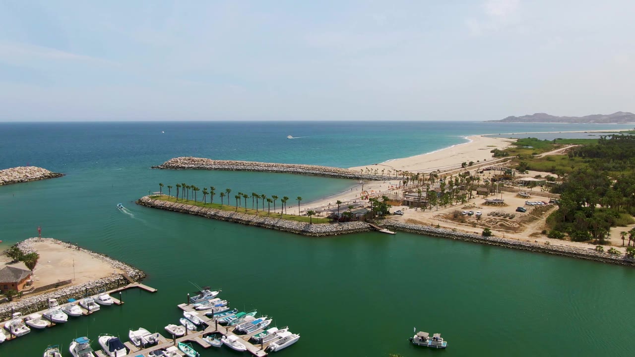 Wide drone frame at Puerto Los Cabos Beach with docked yachts along the lower marina, a small boat near the water, the long rock breakwater, pale sand, blue ocean horizon, and no visible people.
