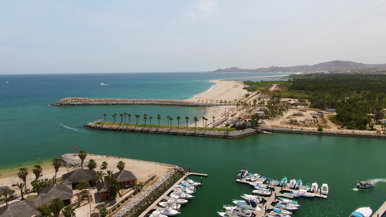 Aerial frame over Puerto Los Cabos Beach with more palm shelters visible near the lower left, docked boats along the marina, green water, long rock breakwaters, sandy beach, blue ocean, and no visible people.