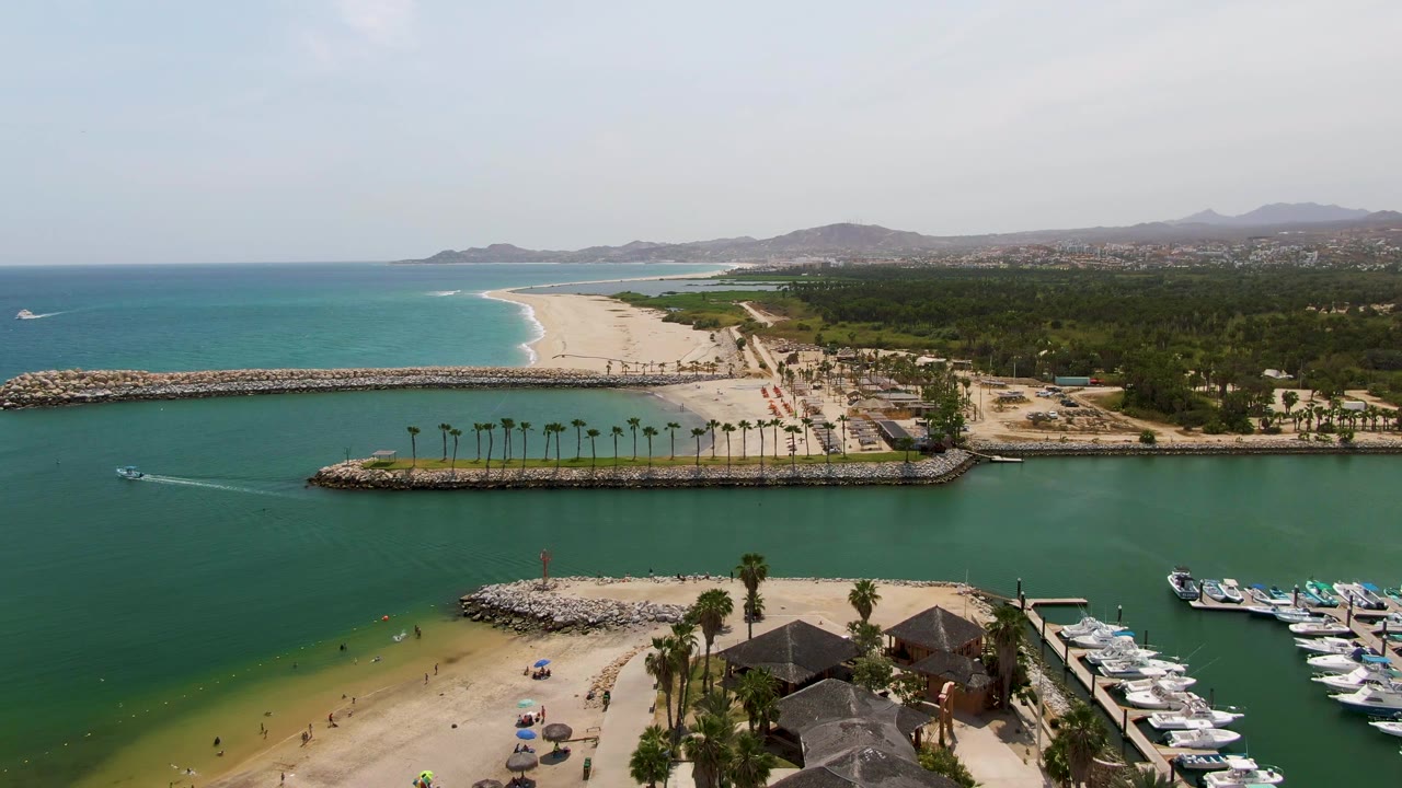 At 0:00, Puerto Los Cabos Beach appears from above with green harbor water, a small boat wake, sandy shore, palm trees, marina boats, breakwaters, distant hills, and tiny visible beachgoers.