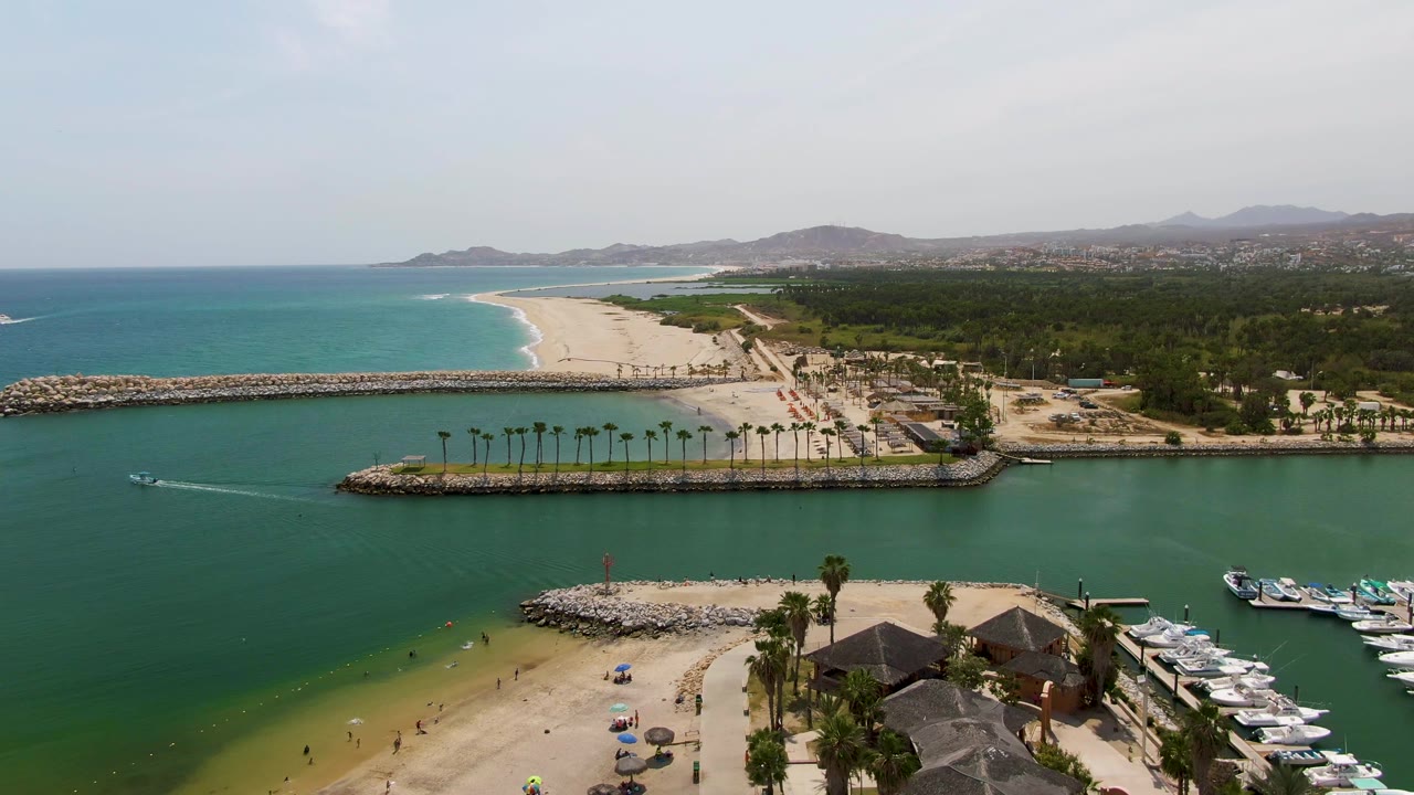Wide aerial harbor view with boat wake, sandy beach, breakwater, palms, and marina slips.