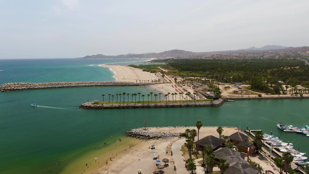 At 0:02, Puerto Los Cabos Beach is framed with the breakwater cutting across the middle, teal ocean beyond the sand, green harbor water below, docked boats at right, and small people on shore.