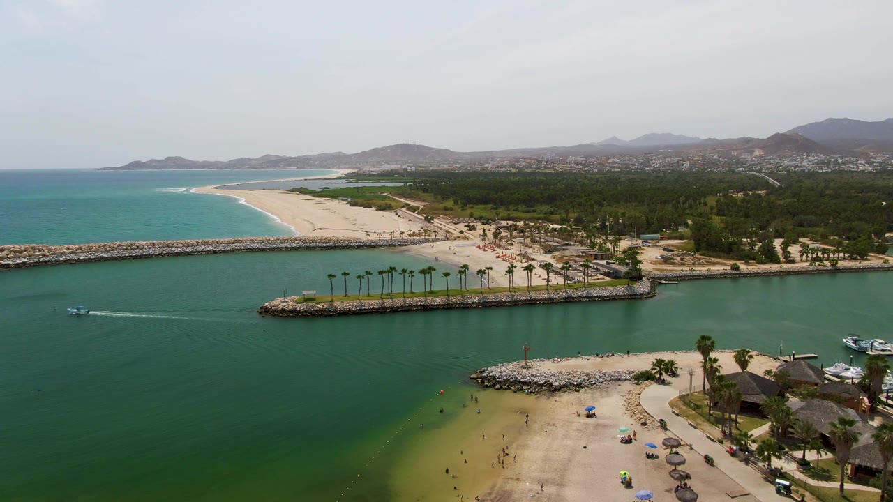 Aerial view of Puerto Los Cabos Beach with green harbor water in front, a white boat wake at left, sandy shore with umbrellas below, rocky breakwaters, turquoise ocean, palms, and hazy hills beyond.