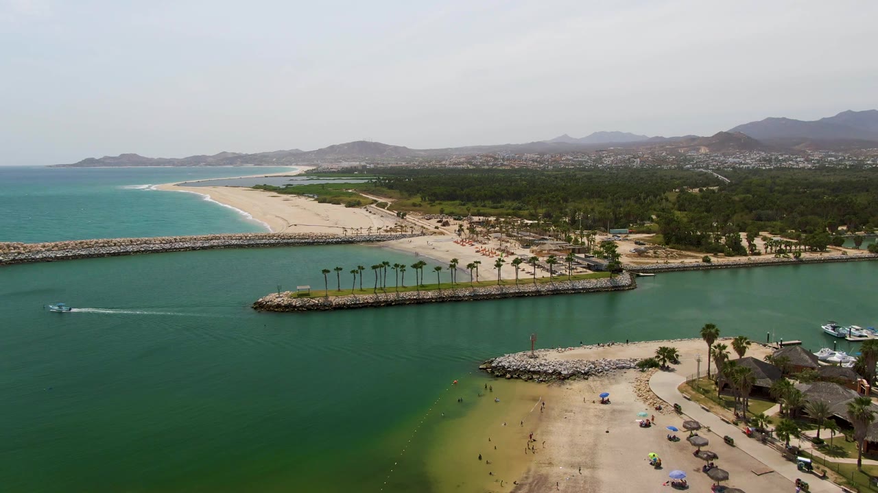Wide aerial of green harbor water, sandy beach, palms, breakwaters, and a boat wake.