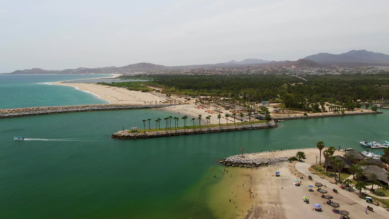 A drone view over Puerto Los Cabos Beach shows the wake cutting through green harbor water, umbrellas and tiny beachgoers along the sand below, a rock breakwater ahead, blue ocean to the left, and low hills in haze.