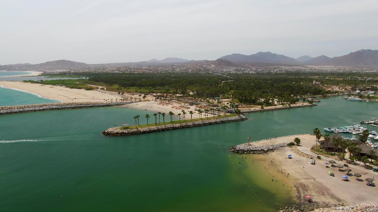 Wide aerial frame over Puerto Los Cabos Beach showing green inlet water in the foreground, a sandy beach and palm-lined breakwater in the midground, marina boats at the right edge, distant mountains, and no visible people.