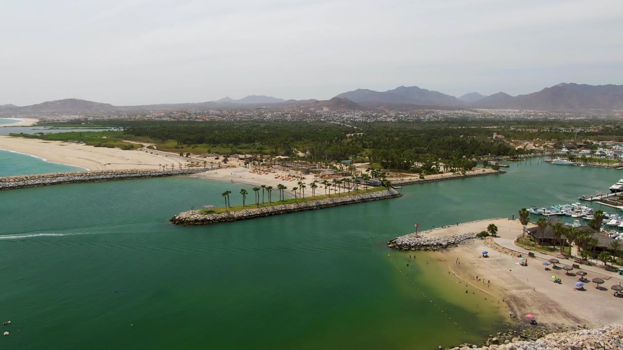 Aerial view at Puerto Los Cabos Beach with the palm-lined breakwater centered, green inlet water below, sandy shore behind it, marina docks and boats to the right, hazy mountains on the horizon, and no visible people.