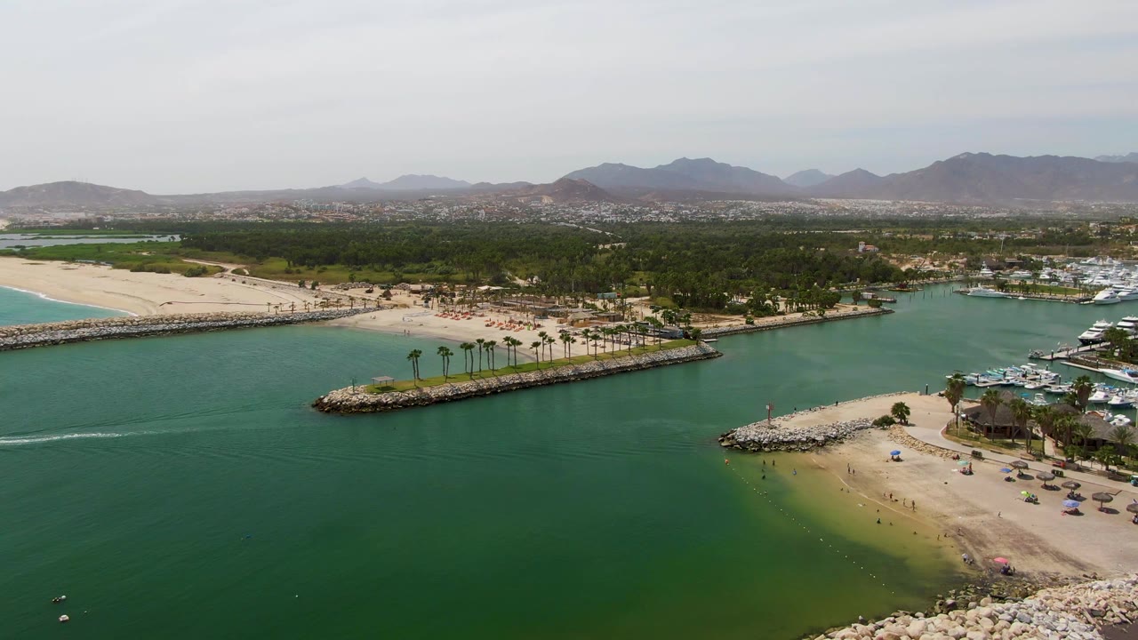 Puerto Los Cabos Beach aerial frame looking across broad green inlet water toward a sandy beach and long breakwater, with marina boats clustered on the right, low mountains in the background, and no visible people.